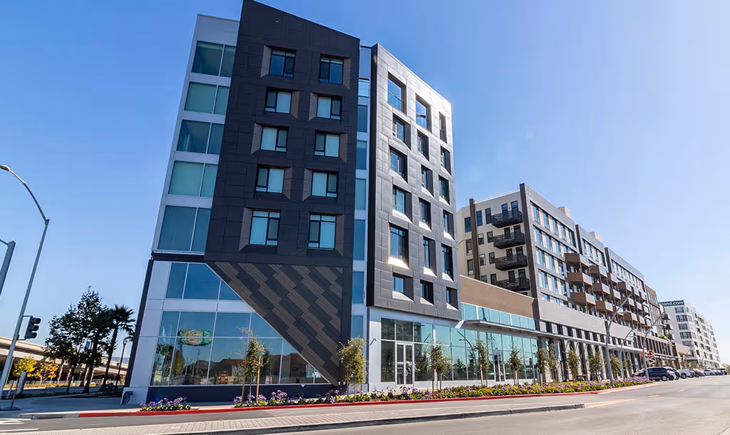 Modern urban resiidential complex with geometric facade, large windows, and colorful flowerbeds. Bright, sunny day enhances the building's sleek design.