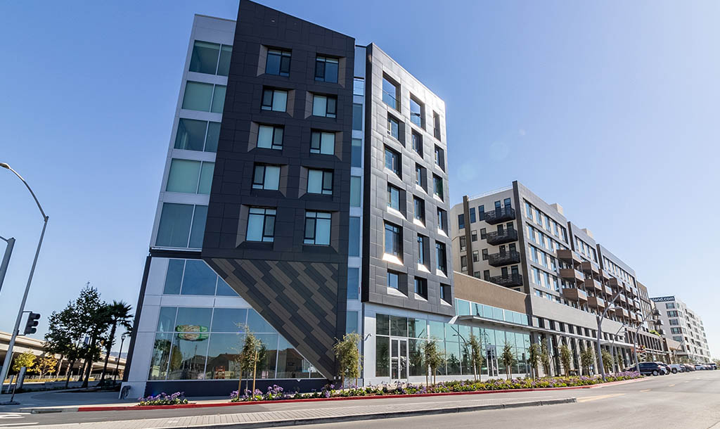Modern urban resiidential complex with geometric facade, large windows, and colorful flowerbeds. Bright, sunny day enhances the building's sleek design.