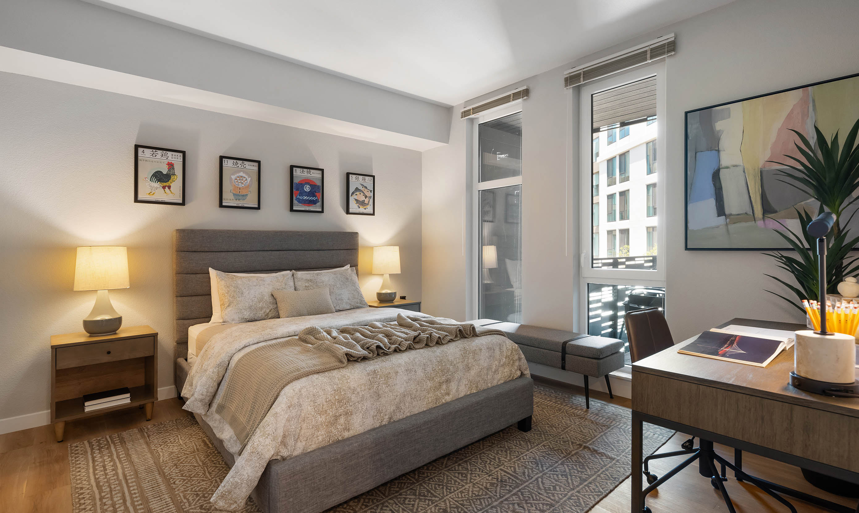 Modern bedroom with warm, neutral tones featuring a padded gray upholstered bed with beige bedding, textured throw blanket, and matching pillows. Two wooden nightstands with ceramic lamps flank the bed. Above the headboard, five framed art prints with Asian-inspired graphics and characters add a cultural touch. A geometric patterned area rug complements the light wood flooring. The room includes a minimalist gray bench by tall windows that allow natural light and showcase a view of a modern apartment building. A sleek wooden desk with a brown leather office chair is positioned in the corner, topped with office supplies, a magazine, and a black desk lamp. A large abstract wall painting and a tall green indoor plant complete the sophisticated, cozy ambiance.