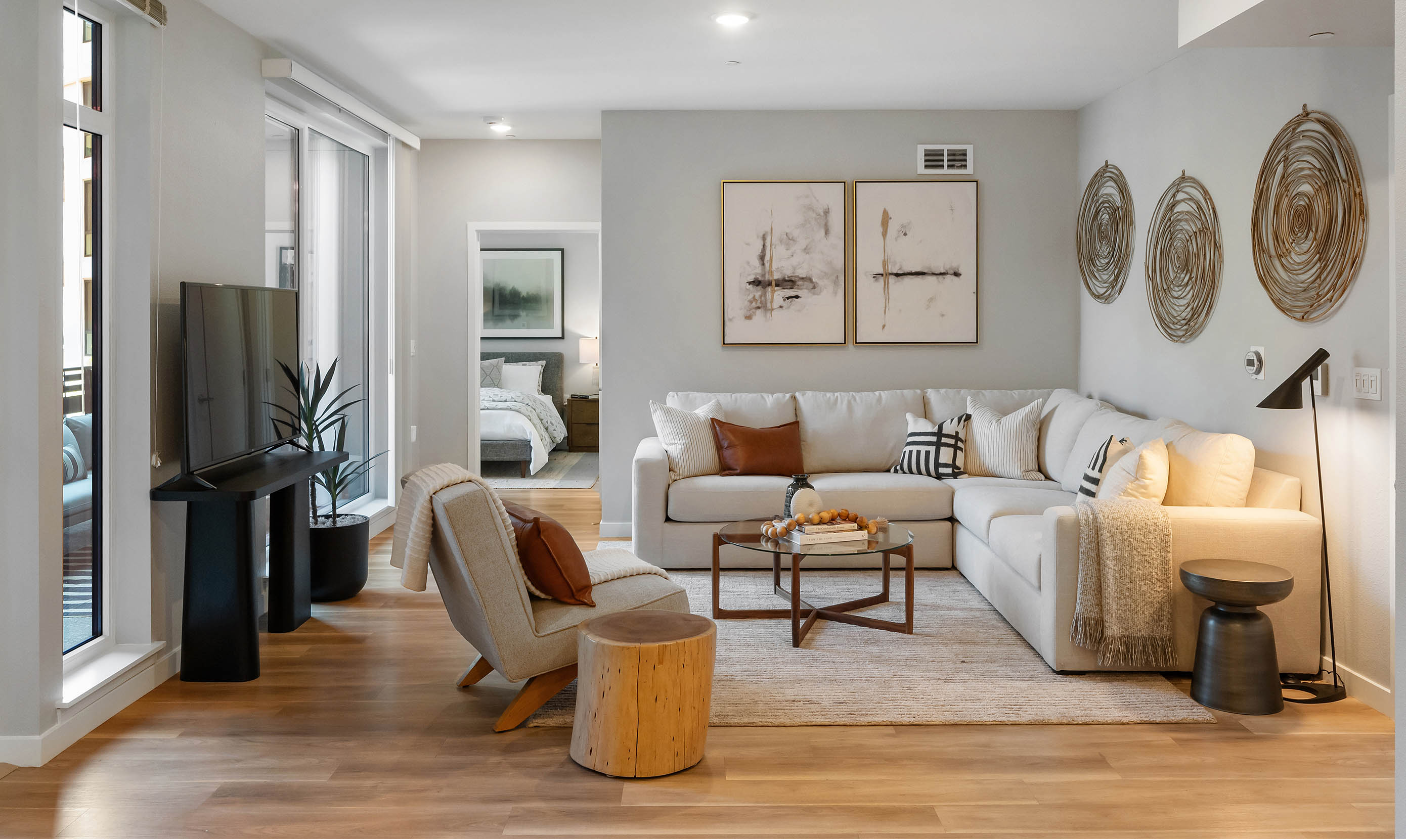 Contemporary open-concept living room with neutral tones featuring a large L-shaped white sectional sofa accented with throw pillows in black, white, and tan. A modern glass coffee table sits on a beige textured area rug atop light wood flooring. Wall decor includes abstract paintings and three large circular wicker wall hangings. A minimalist lounge chair with a tan throw and a small wooden stump side table add texture near a sleek black TV stand with a flat-screen TV. A tall indoor plant in a black pot adds greenery beside floor-to-ceiling windows. A bedroom with a neatly made bed is visible through an open doorway, enhancing the modern, cozy ambiance.