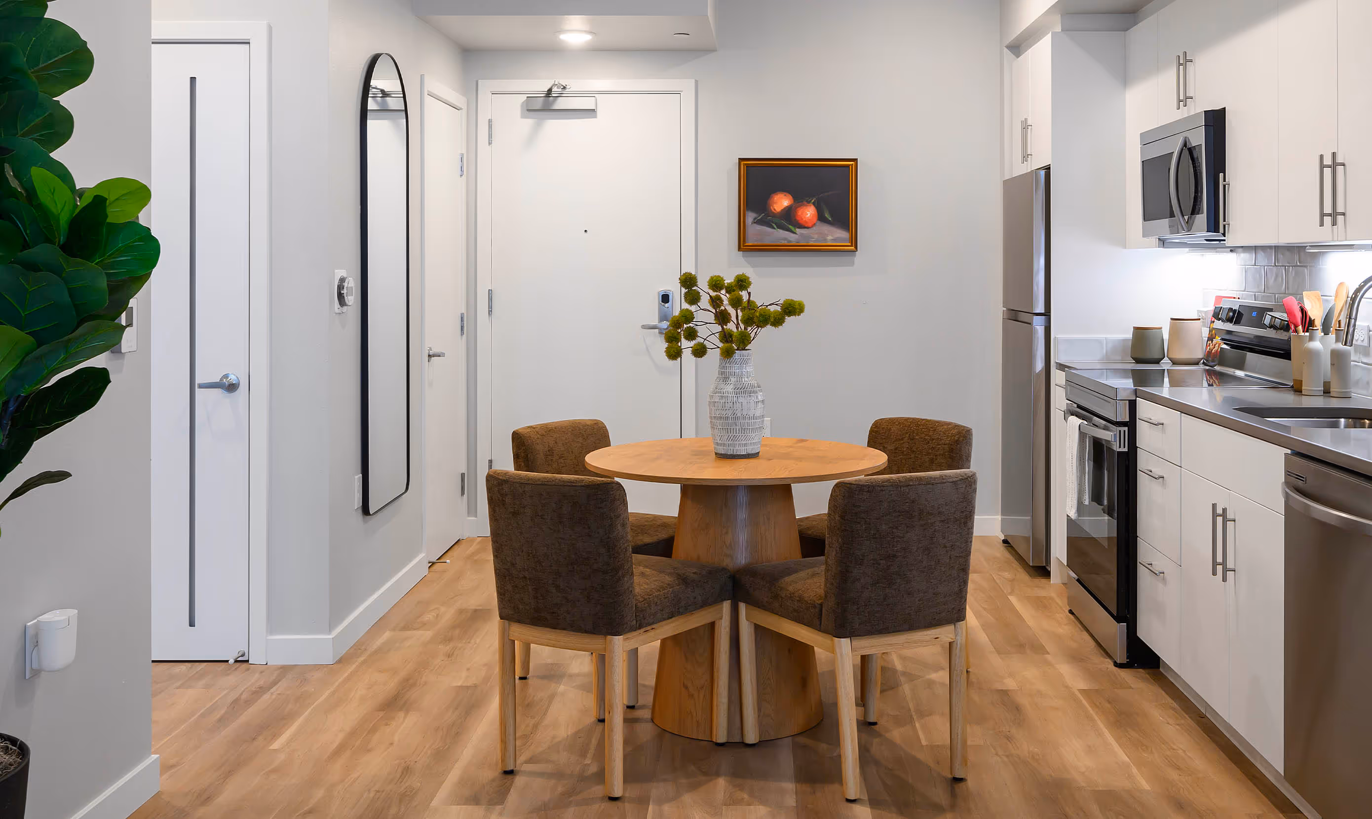 Contemporary kitchen and dining area with minimalist design, featuring a compact round wooden dining table surrounded by four brown upholstered chairs. The kitchen is equipped with sleek white cabinetry, stainless steel appliances including a refrigerator, oven, microwave, and dishwasher, and a glossy gray backsplash. A decorative ceramic vase with green botanicals sits atop the table as a centerpiece. The space is accented with warm wood-tone flooring, a wall-mounted full-length mirror, a framed painting of two red fruits, and a tall potted fiddle-leaf fig plant. Multiple doors lead to adjacent rooms, and recessed lighting provides a clean, modern ambiance.