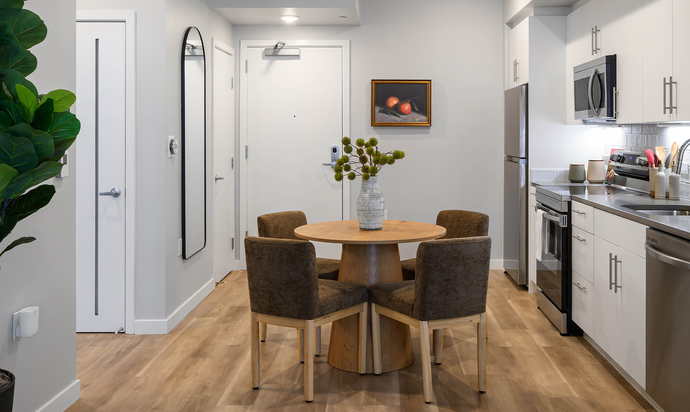 Contemporary kitchen and dining area with minimalist design, featuring a compact round wooden dining table surrounded by four brown upholstered chairs. The kitchen is equipped with sleek white cabinetry, stainless steel appliances including a refrigerator, oven, microwave, and dishwasher, and a glossy gray backsplash. A decorative ceramic vase with green botanicals sits atop the table as a centerpiece. The space is accented with warm wood-tone flooring, a wall-mounted full-length mirror, a framed painting of two red fruits, and a tall potted fiddle-leaf fig plant. Multiple doors lead to adjacent rooms, and recessed lighting provides a clean, modern ambiance.