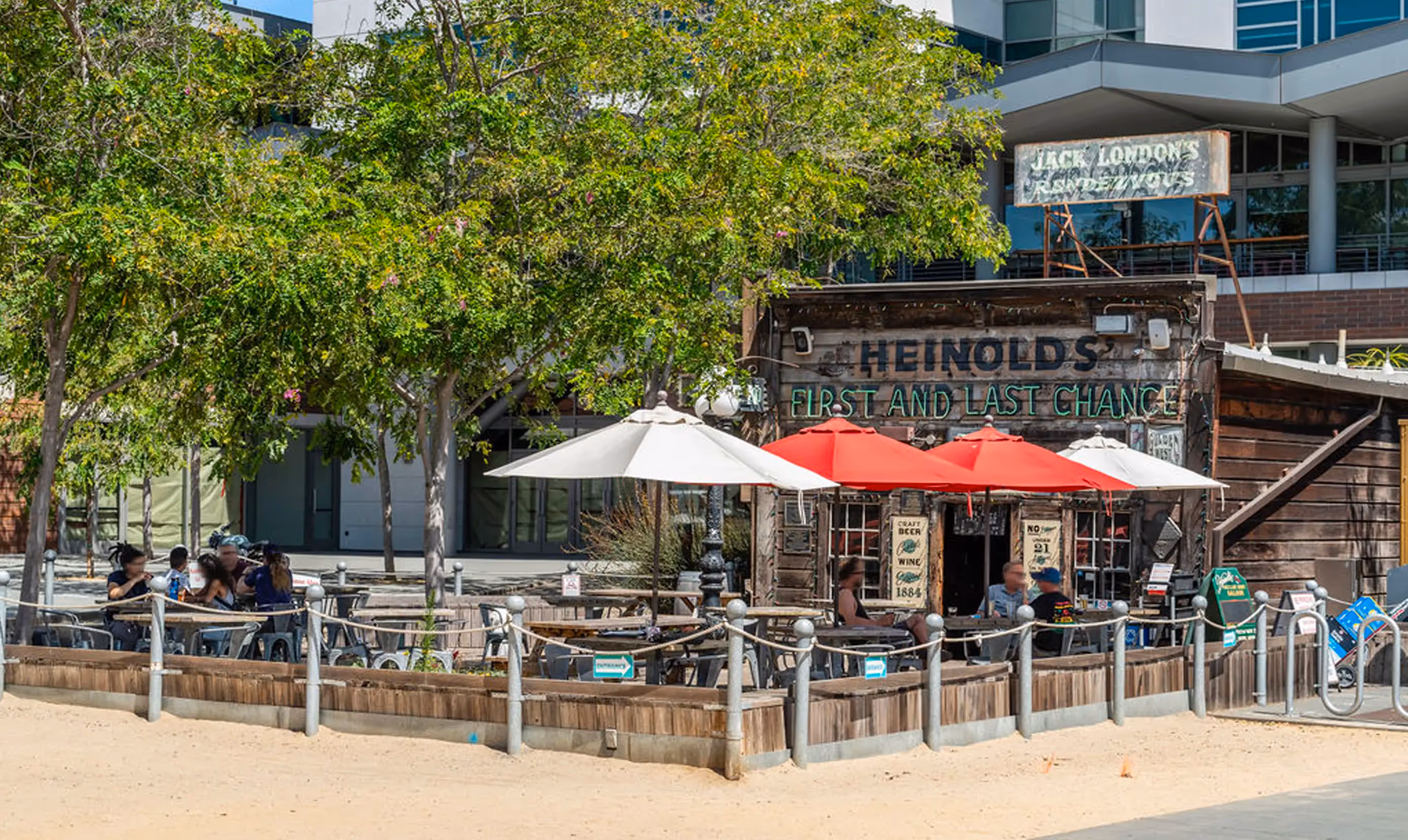 Outdoor view of Heinold’s First and Last Chance Saloon, a historic wooden bar located in Jack London Square, Oakland, California. The rustic exterior features weathered wood siding, vintage signage, and a prominent sign reading “Jack London’s Rendezvous.” Patrons are seated under red and white patio umbrellas at metal tables in the shaded outdoor seating area surrounded by a low wooden barrier. Trees provide natural shade, and a sandy path runs along the edge of the patio. Modern buildings are visible in the background, creating a contrast between the historic saloon and contemporary architecture.