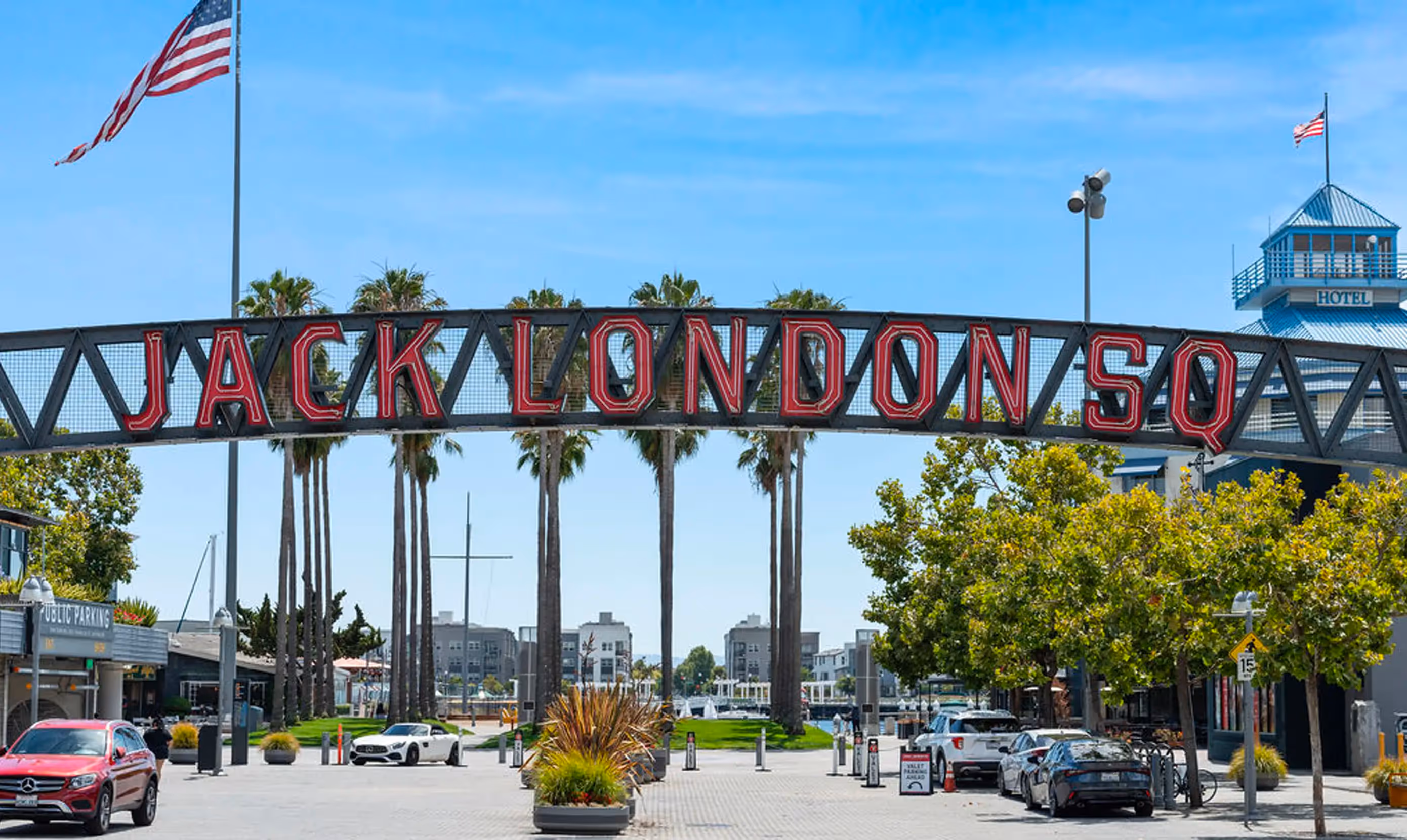 Iconic entrance sign to Jack London Square in Oakland, California, featuring large red neon-style letters mounted on a black truss archway. The area is lined with tall palm trees and has a mix of parked cars, a valet stand, and visible signage for public parking. In the background, there are modern waterfront buildings and a hotel with a blue rooftop. Two American flags are flying, one prominently on a tall flagpole to the left. The sky is clear and blue, reflecting a sunny day at this popular waterfront destination.