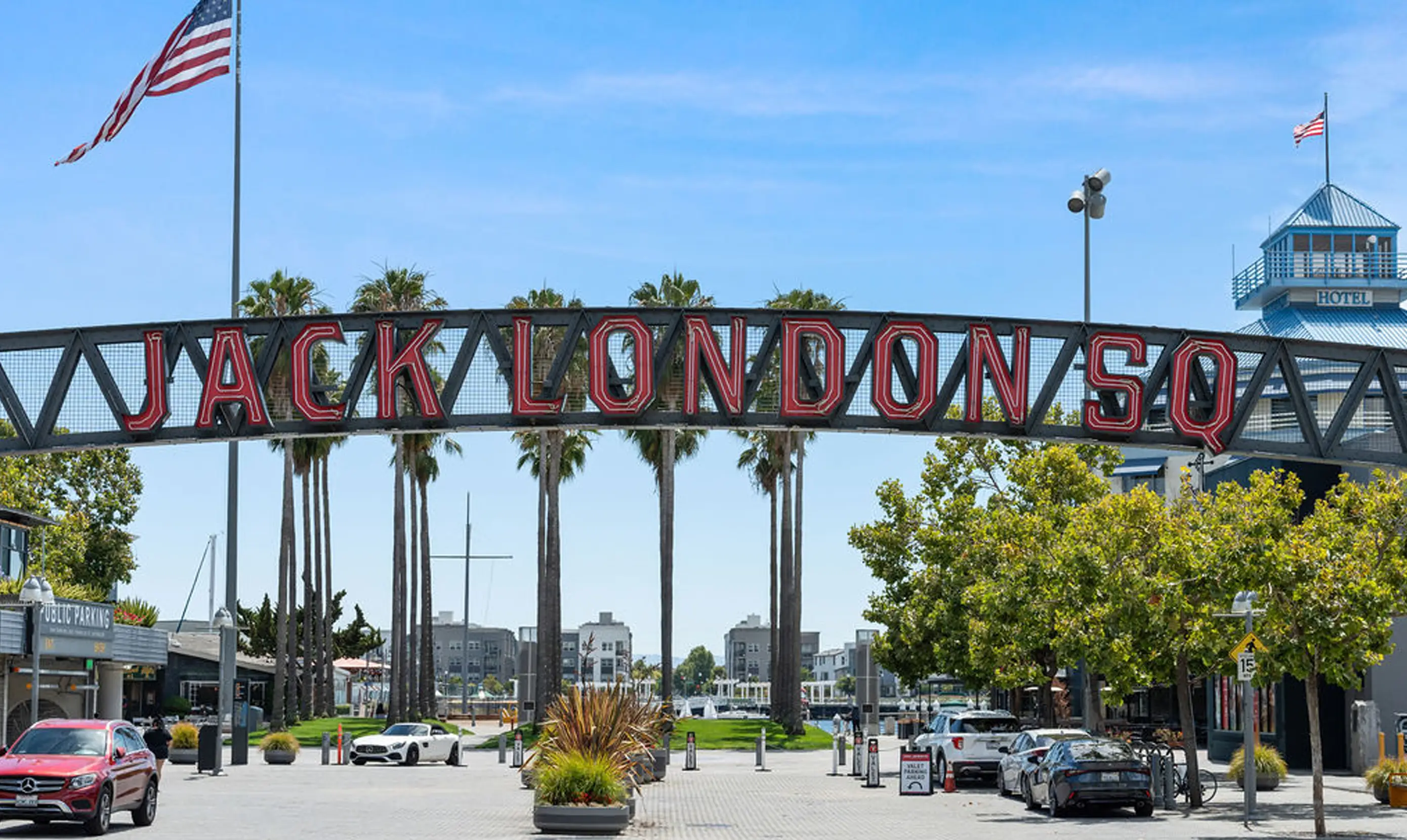 Iconic entrance sign to Jack London Square in Oakland, California, featuring large red neon-style letters mounted on a black truss archway. The area is lined with tall palm trees and has a mix of parked cars, a valet stand, and visible signage for public parking. In the background, there are modern waterfront buildings and a hotel with a blue rooftop. Two American flags are flying, one prominently on a tall flagpole to the left. The sky is clear and blue, reflecting a sunny day at this popular waterfront destination.