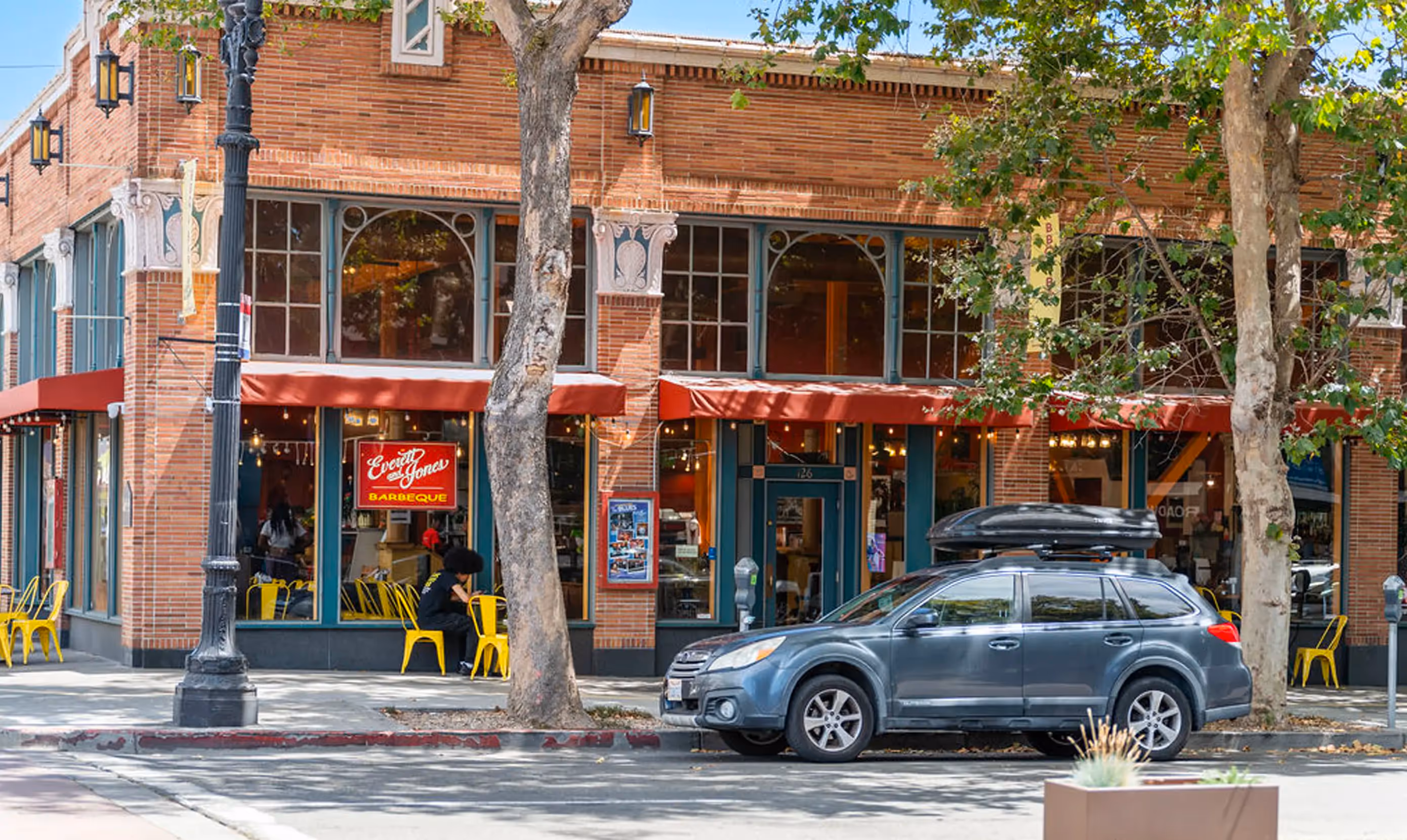 Street view of Everett & Jones Barbeque restaurant in Oakland, California, located in a historic red-brick building with large arched windows and red awnings. Bright yellow metal chairs line the sidewalk patio seating area. A customer sits outside under the shade of mature trees, and a gray SUV is parked at the curb. The restaurant's glowing red sign reading “Everett & Jones Barbeque” is prominently displayed in the window.