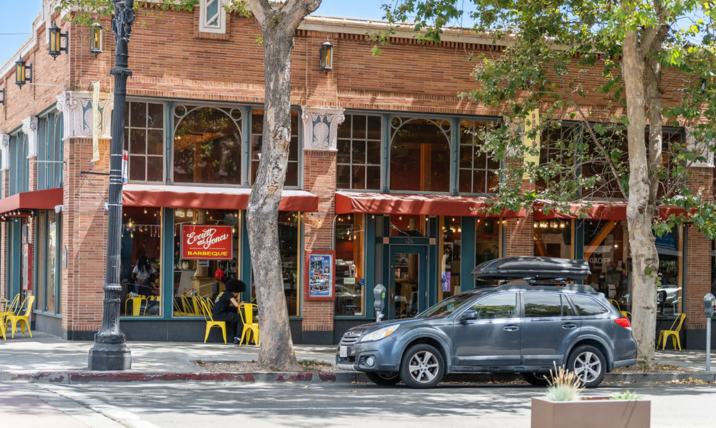Street view of Everett & Jones Barbeque restaurant in Oakland, California, located in a historic red-brick building with large arched windows and red awnings. Bright yellow metal chairs line the sidewalk patio seating area. A customer sits outside under the shade of mature trees, and a gray SUV is parked at the curb. The restaurant's glowing red sign reading “Everett & Jones Barbeque” is prominently displayed in the window.