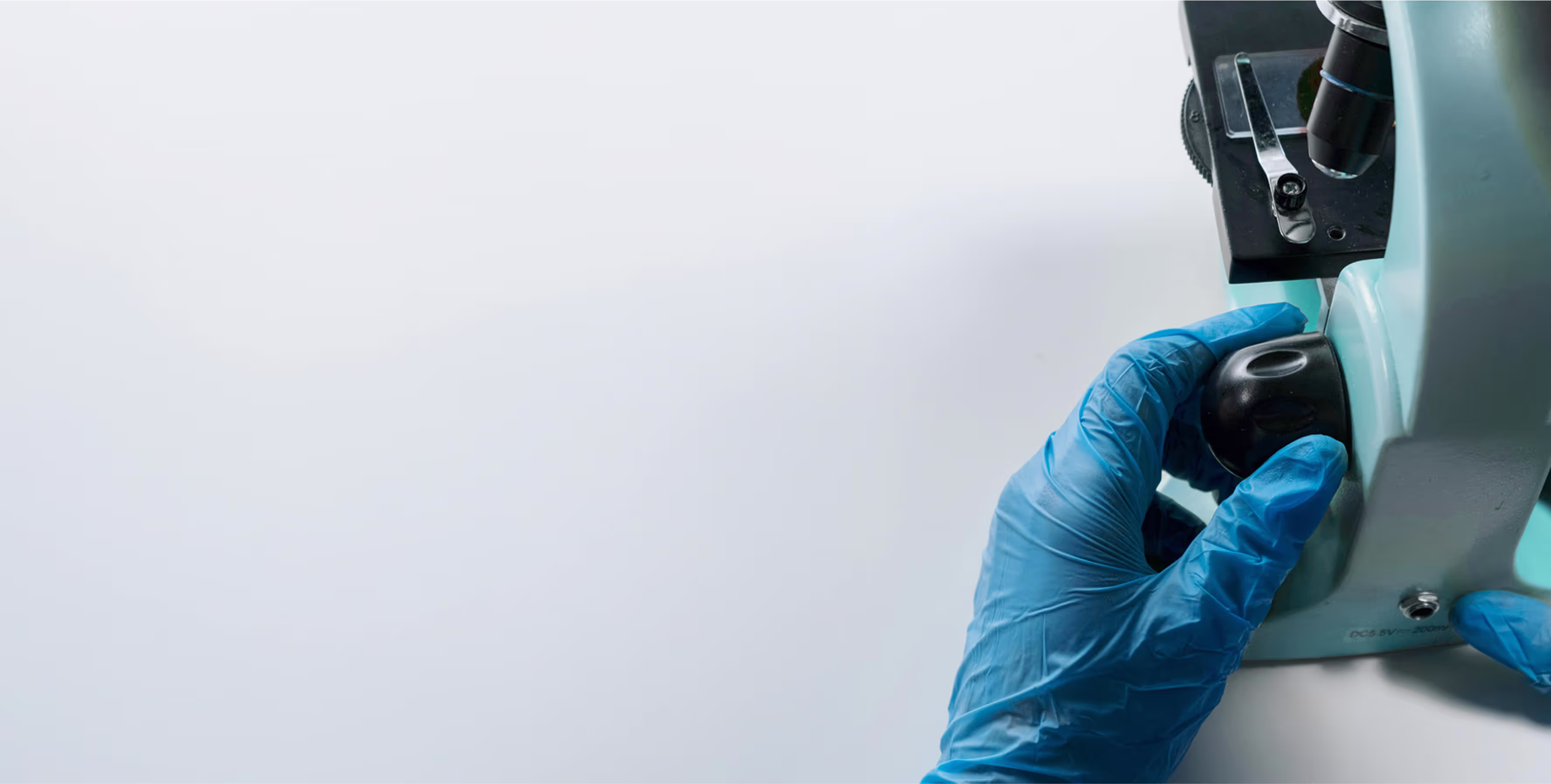 A person wearing blue gloves adjusts a microscope in a laboratory setting. The focus is on the gloved hands turning a knob on the microscope, with a clean, white background emphasizing the equipment.