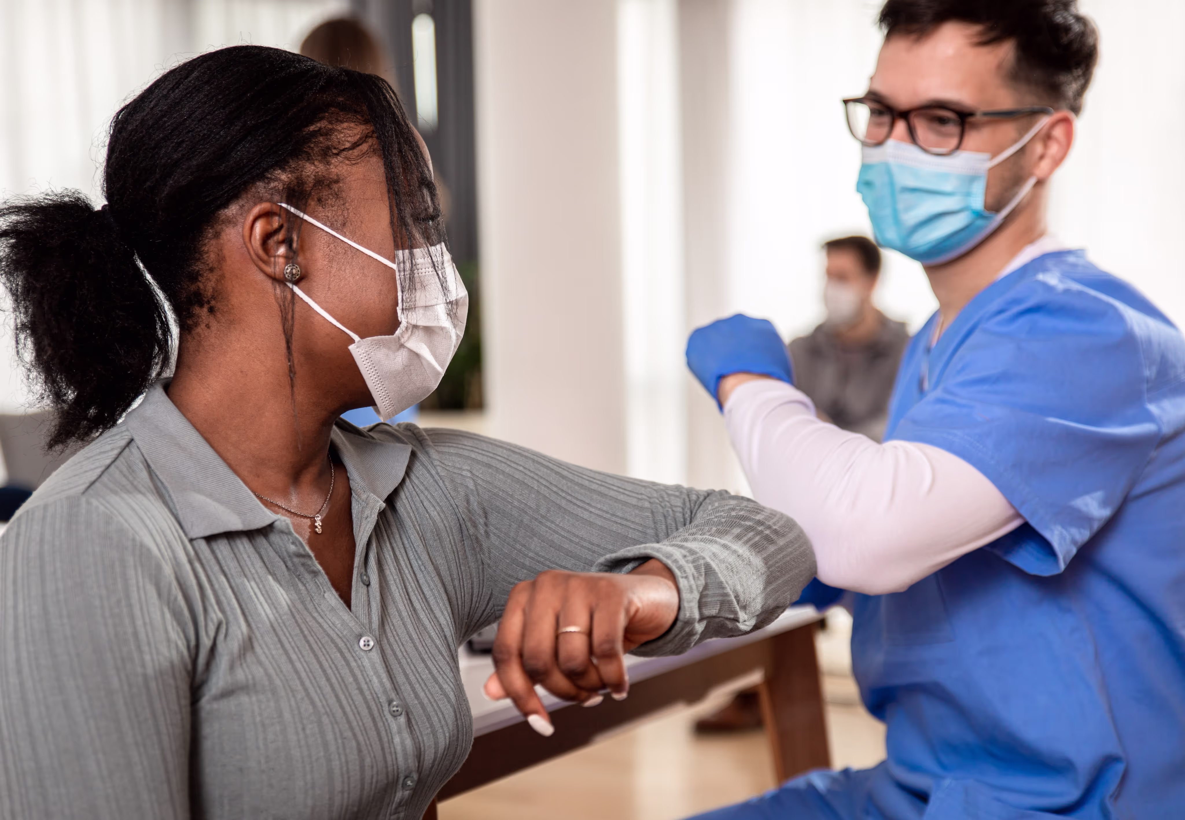 A medical professional in blue scrubs and gloves checks a patient's arm in a clinical setting. Both are wearing face masks, and the professional gently holds the patient's wrist. The background shows a bright room with another person in a white coat observing.