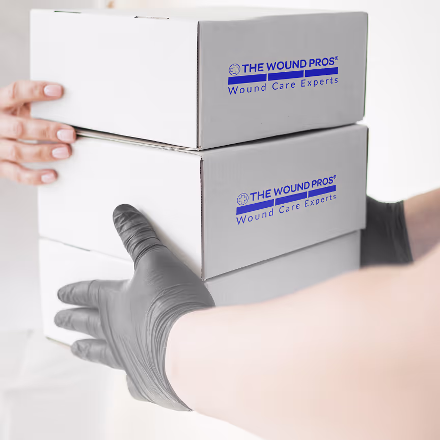 A person wearing a gray glove holds two white boxes stacked together, each labeled "The Wound Pros Wound Care Experts" in blue text. The focus is on the hands and boxes, with a clean, neutral background.
