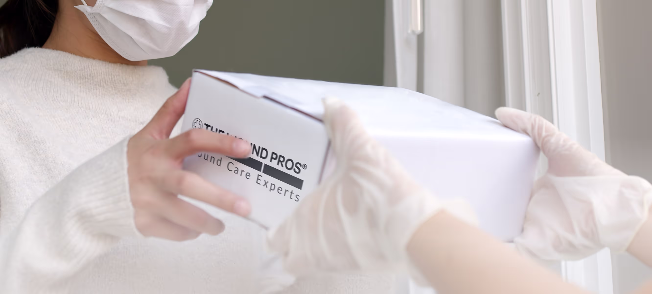 A person wearing a face mask and gloves hands over a white package labeled "The Wound Pros Wound Care Experts" to another gloved individual. The exchange occurs indoors near a window with curtains, suggesting a safe and controlled delivery process.
