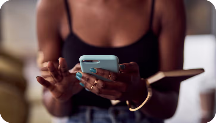 A person wearing a black top and gold bracelets uses a light blue smartphone, holding a notebook in the other hand. The scene is set against a blurred indoor background, suggesting a casual or work-related setting.