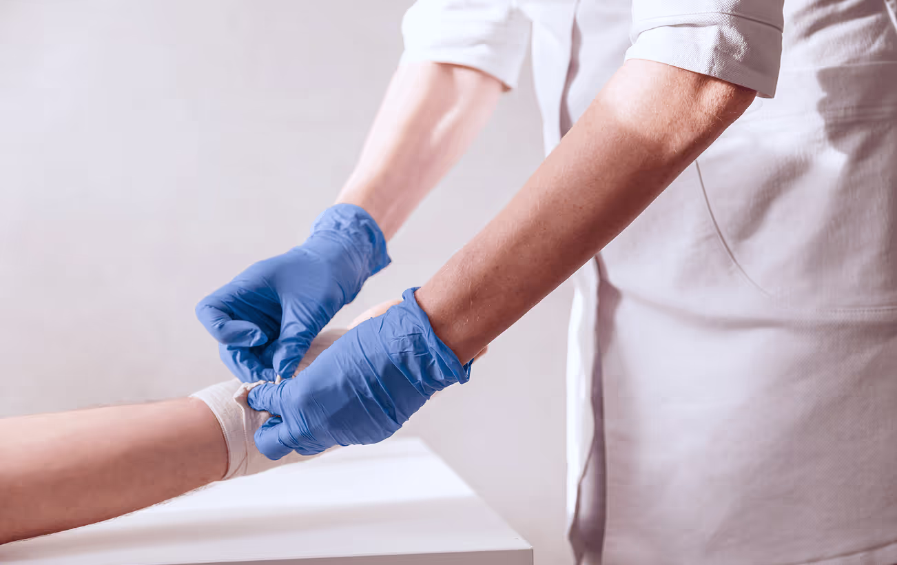 A healthcare professional wearing blue gloves is bandaging a patient's arm in a clinical setting.