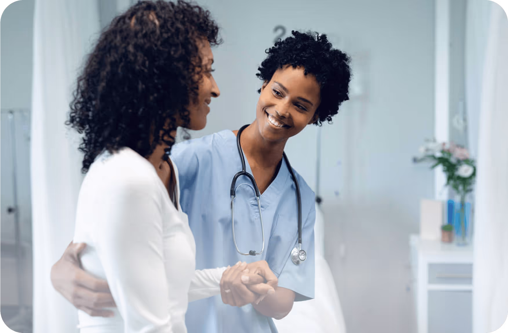 A nurse in blue scrubs with a stethoscope around her neck holds the hand of a patient in a white top, offering comfort in a clinical room. The background includes medical furniture and a vase with flowers, creating a calm and supportive atmosphere.