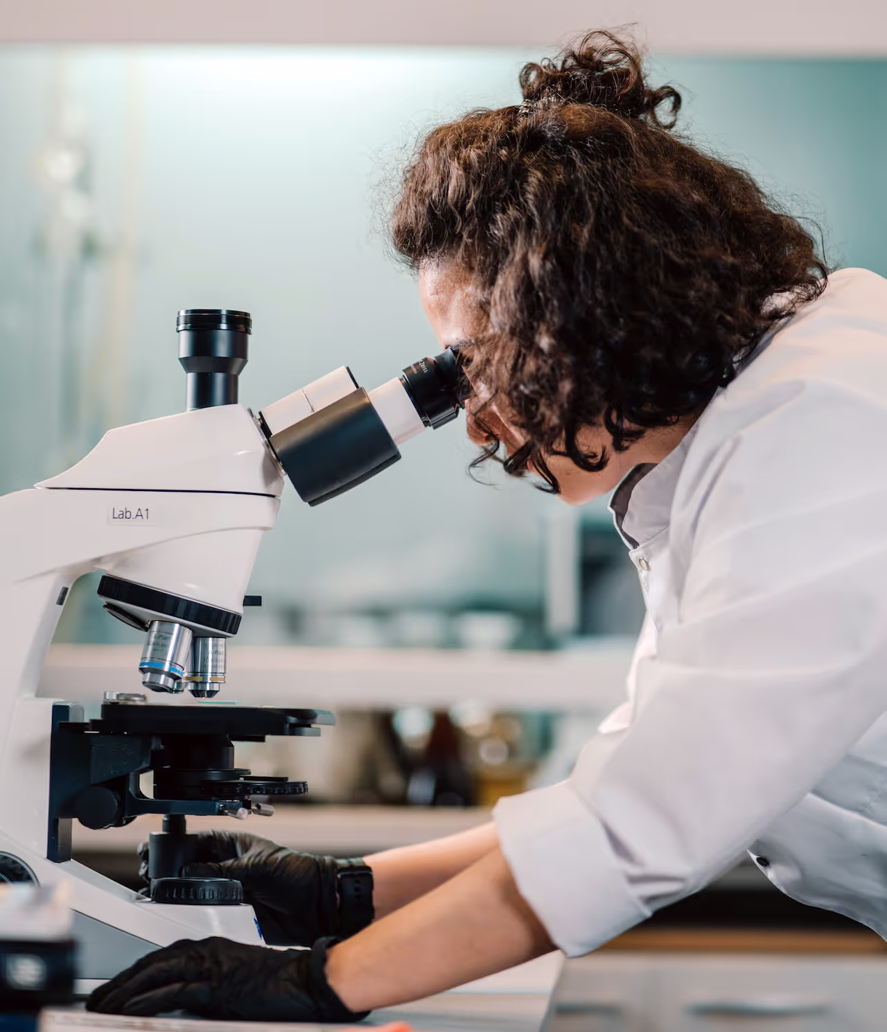 A scientist in a white lab coat and black gloves uses a microscope labeled "LAB A1" in a laboratory setting. The background shows a clean, modern lab environment with blurred equipment and shelves.