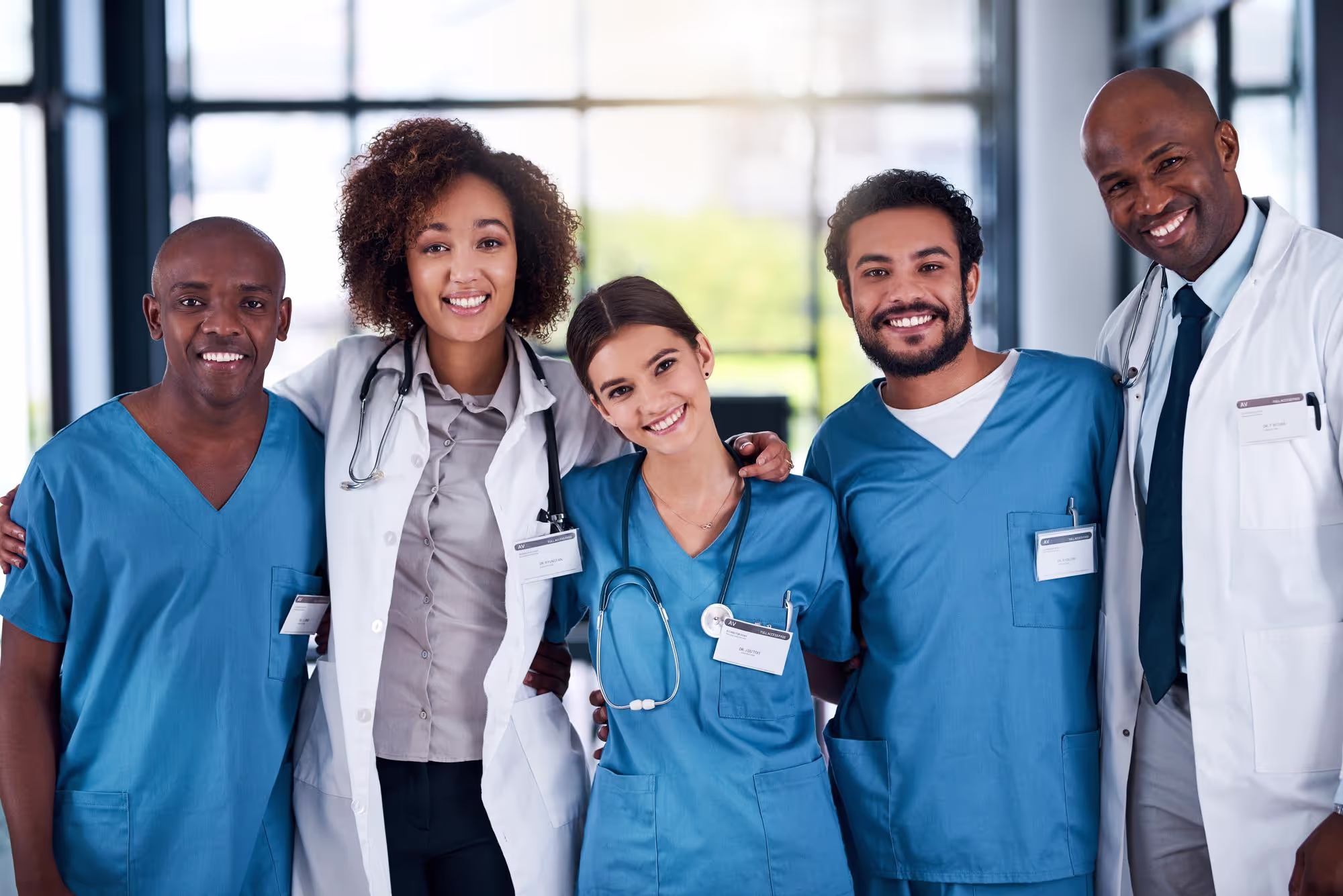 A group of five smiling medical professionals posing together in a bright room with large windows. Four are wearing blue scrubs, and one is in a white coat with a stethoscope. They stand close, with a positive and collaborative atmosphere.