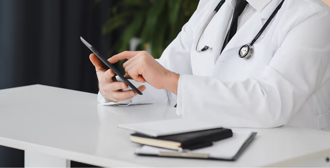 A doctor in a white coat with a stethoscope around their neck uses a smartphone at a desk. The desk holds a few notebooks, and the background features a blurred green plant and a curtain, indicating a clinical office setting.