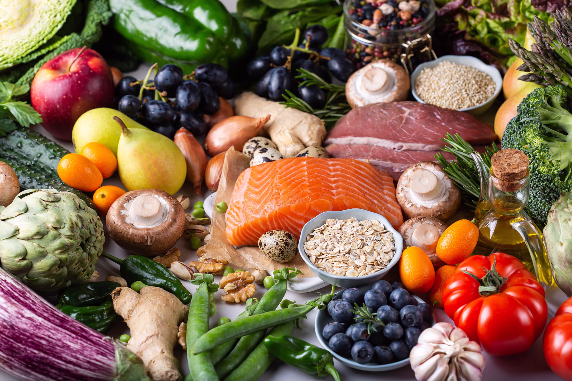 A vibrant assortment of healthy foods on a table, including salmon, broccoli, tomatoes, blueberries, oranges, mushrooms, quinoa, nuts, and olive oil. The colorful spread highlights nutritious ingredients beneficial for wound healing, arranged against a light background.