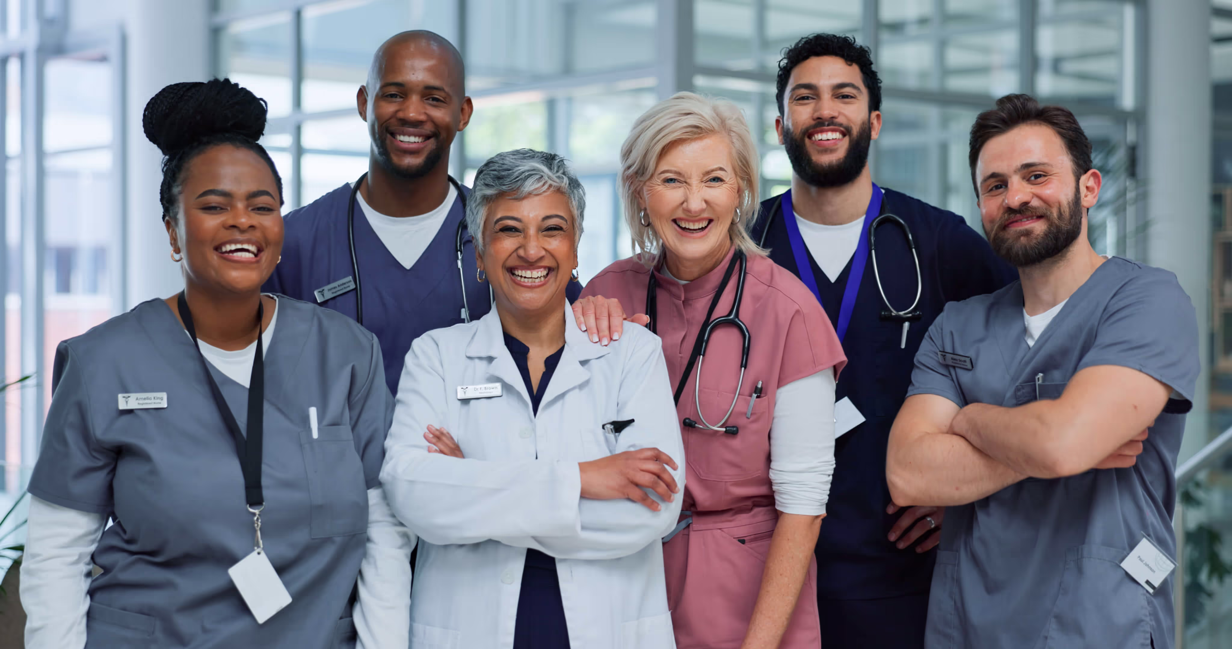 A diverse group of six smiling medical professionals posing together in a bright room with large windows. They wear a mix of blue scrubs, a white coat, and pink scrubs, standing close with a sense of teamwork and positivity.