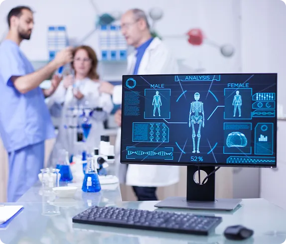 Healthcare professionals in a lab reviewing digital medical scans and data on a computer monitor, with medical equipment in the background.