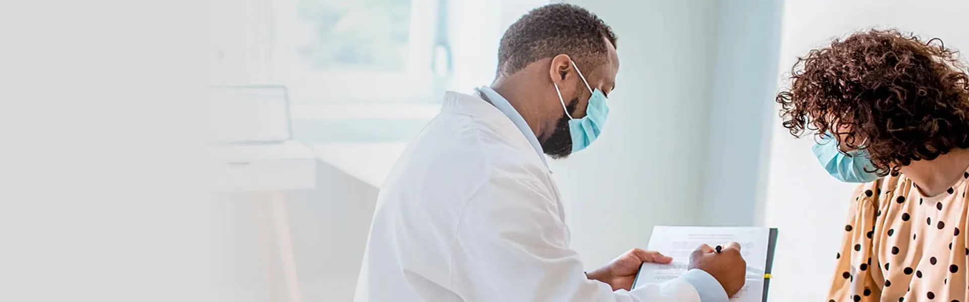 A healthcare professional in a white coat and mask consulting with a patient holding a tablet in a clinical setting.