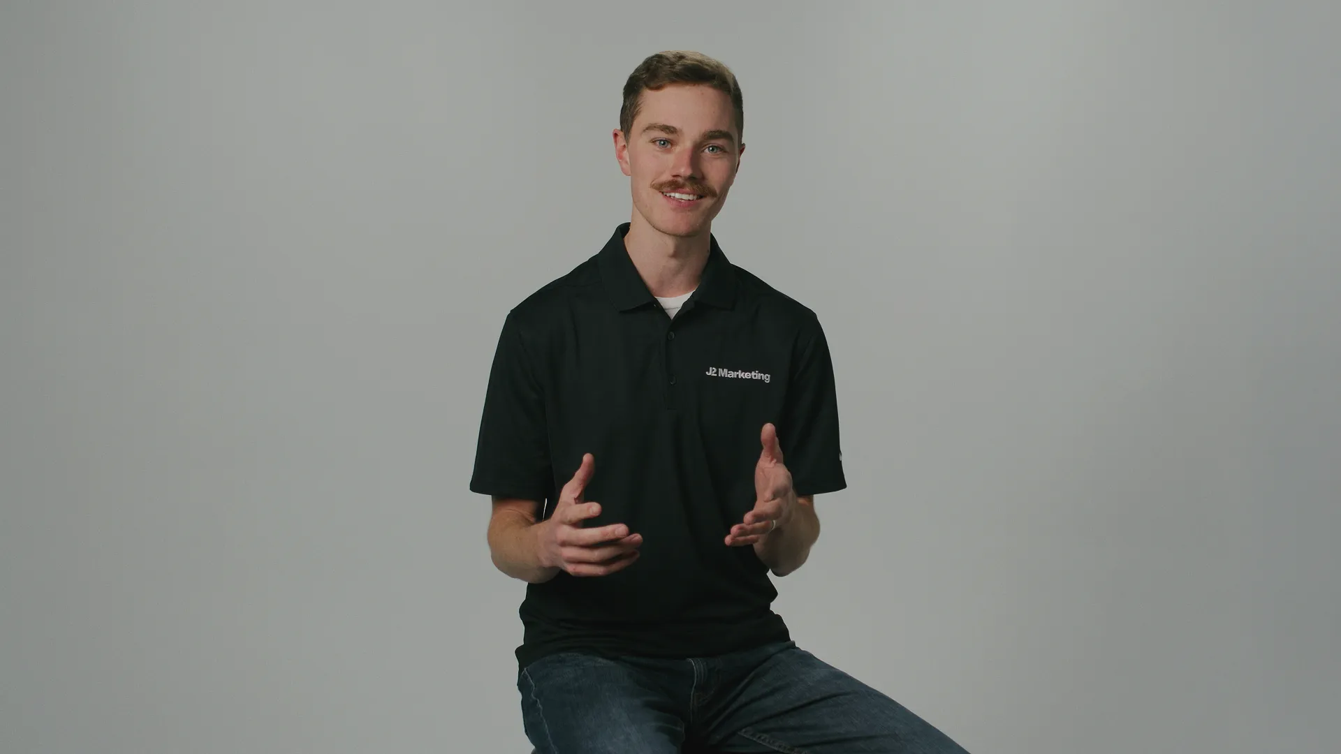 Young man with short brown hair and mustache wearing a black J2 Marketing polo shirt, seated against a plain gray background, gesturing with his hands while smiling.