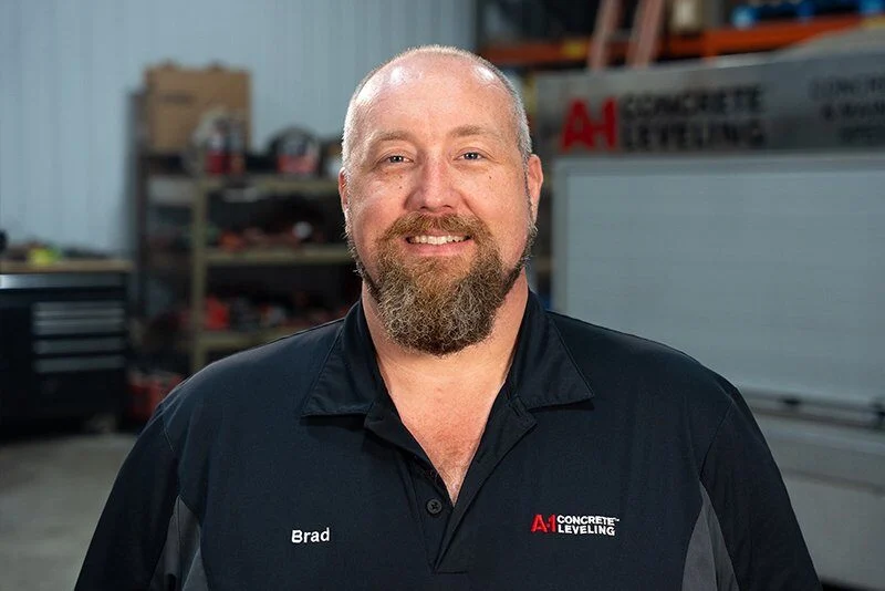 Smiling man with a beard and shaved head wearing a black A1 Concrete Leveling shirt in a workshop.