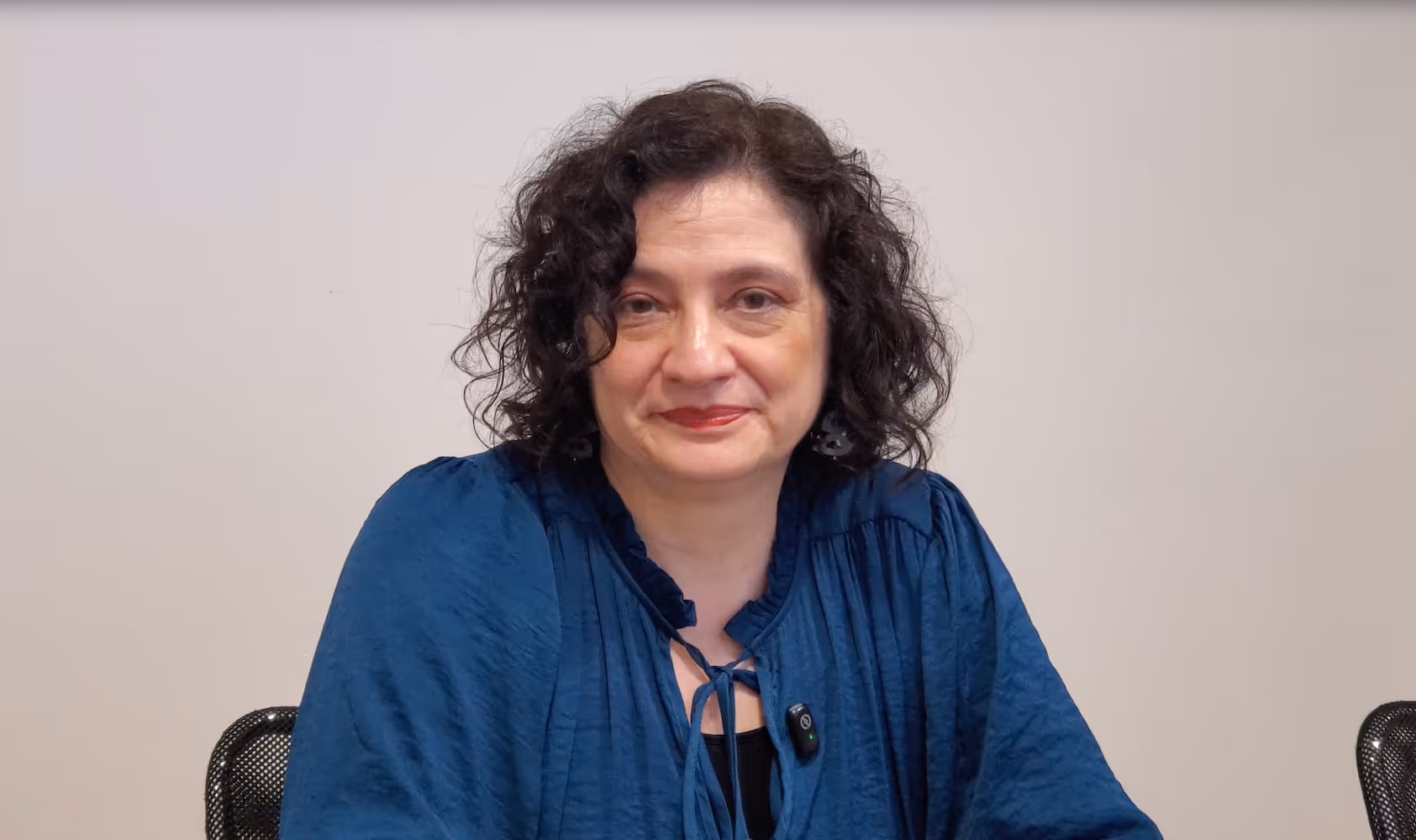Woman with curly dark hair wearing a blue blouse, seated against a plain light background.