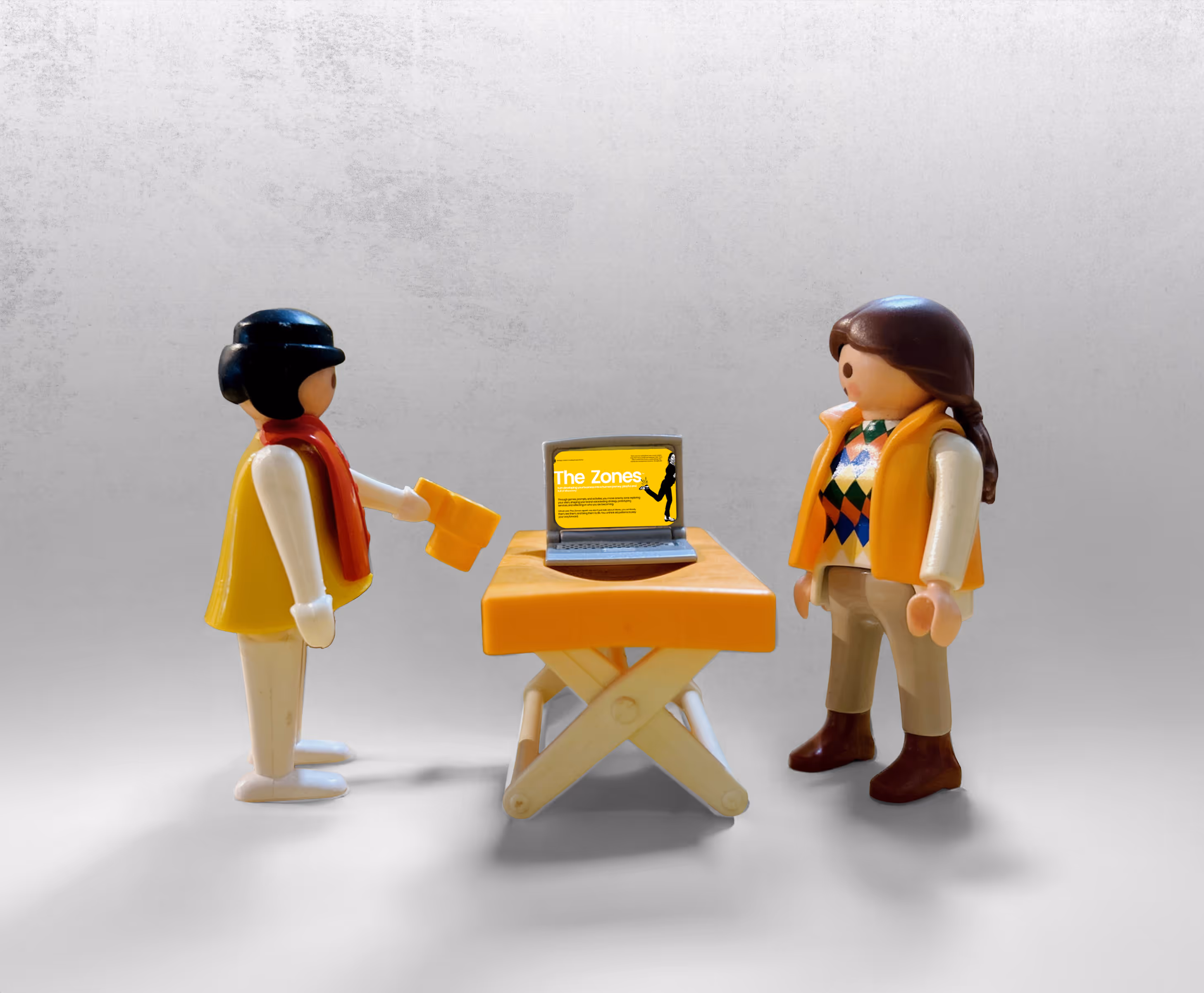 Two LEGO figurines of women standing by a toy table, facing each other, with a toy laptop on the table showing a presentation of the game ‘Zones.’