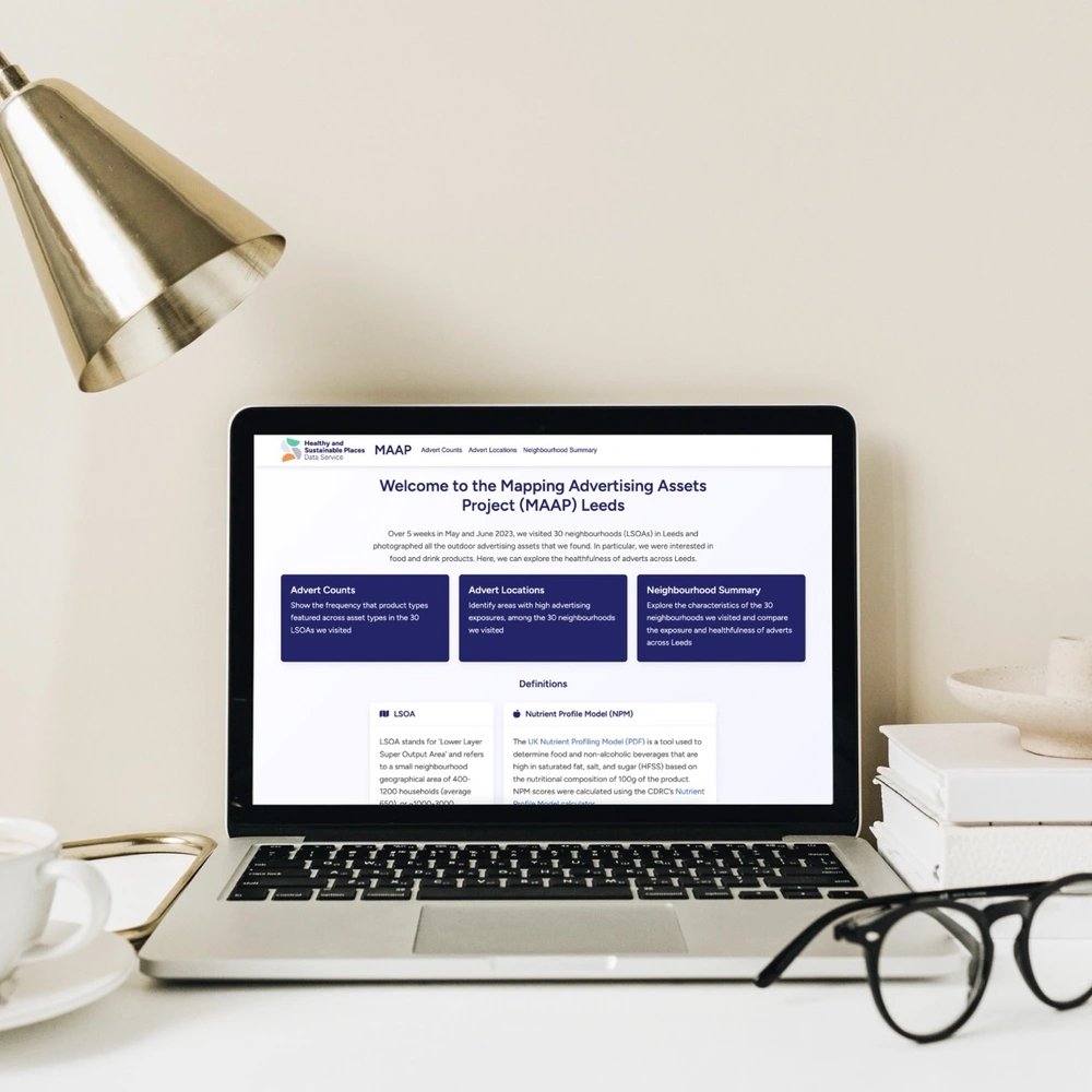 Laptop on desk displaying the Mapping Advertising Assets Project (MAAP) Leeds website, surrounded by a gold lamp, books, glasses, and a cup on a saucer.