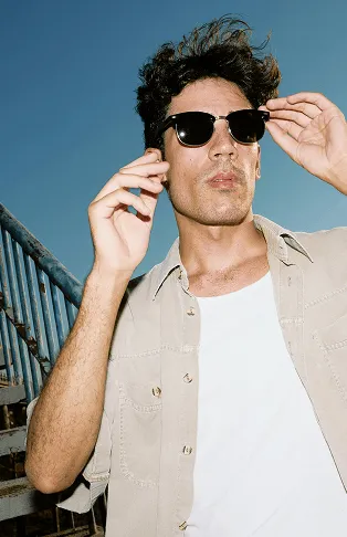 Homme portant des lunettes de soleil noires, chemise beige ouverte sur un t-shirt blanc, posant devant un escalier extérieur sous un ciel bleu.