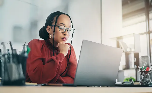 Jeune femme portant des lunettes, réfléchissant devant un ordinateur portable dans un bureau lumineux.