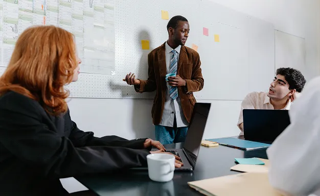 Un homme debout présente des idées avec une carte sur un tableau blanc, tandis que deux collègues à table l'écoutent attentivement, avec des ordinateurs portables devant eux.
