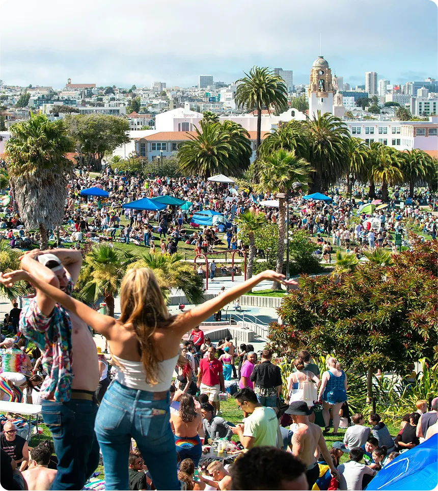 Multitud de personas disfrutando de un evento al aire libre en un parque urbano con palmeras y edificios al fondo.