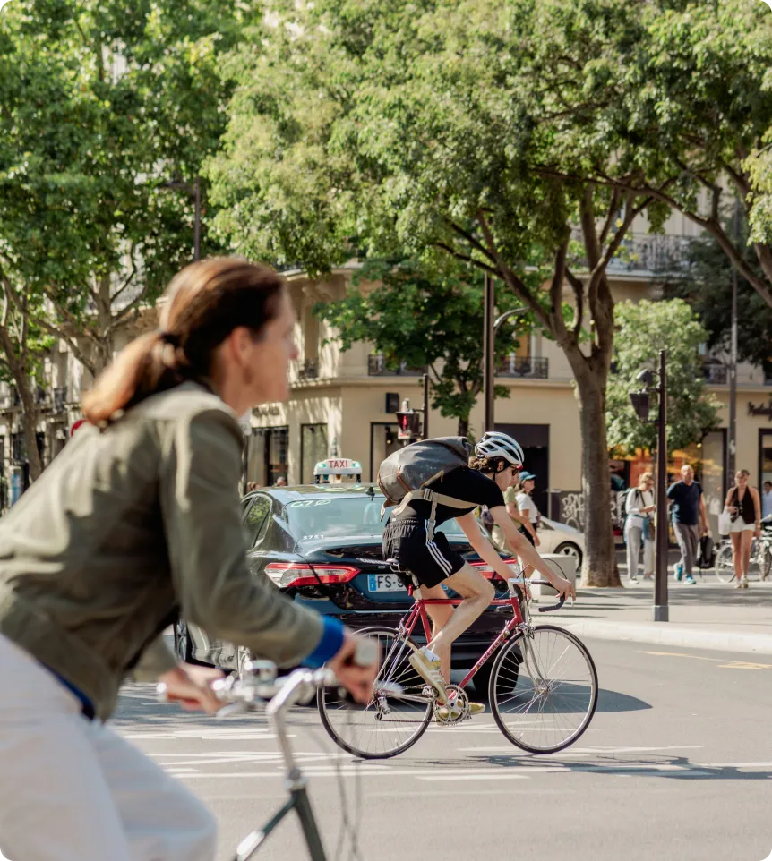 A man on a bicycle wearing a helmet and carrying a backpack rides ahead of a black cab on a busy urban street with passers-by and trees.