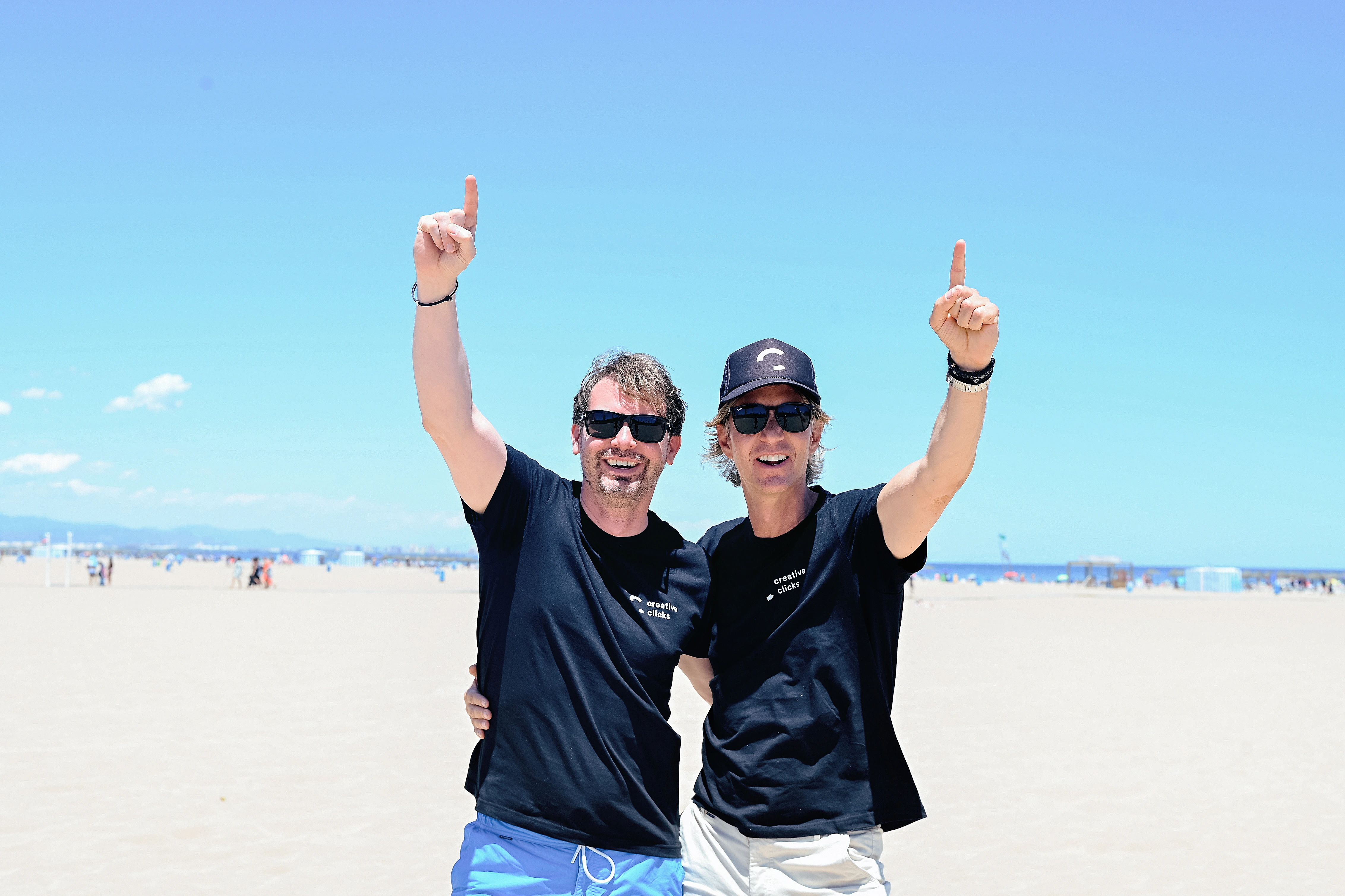 Two men wearing sunglasses and matching black T-shirts standing on a sunny beach, smiling with their arms raised and pointing upward.
