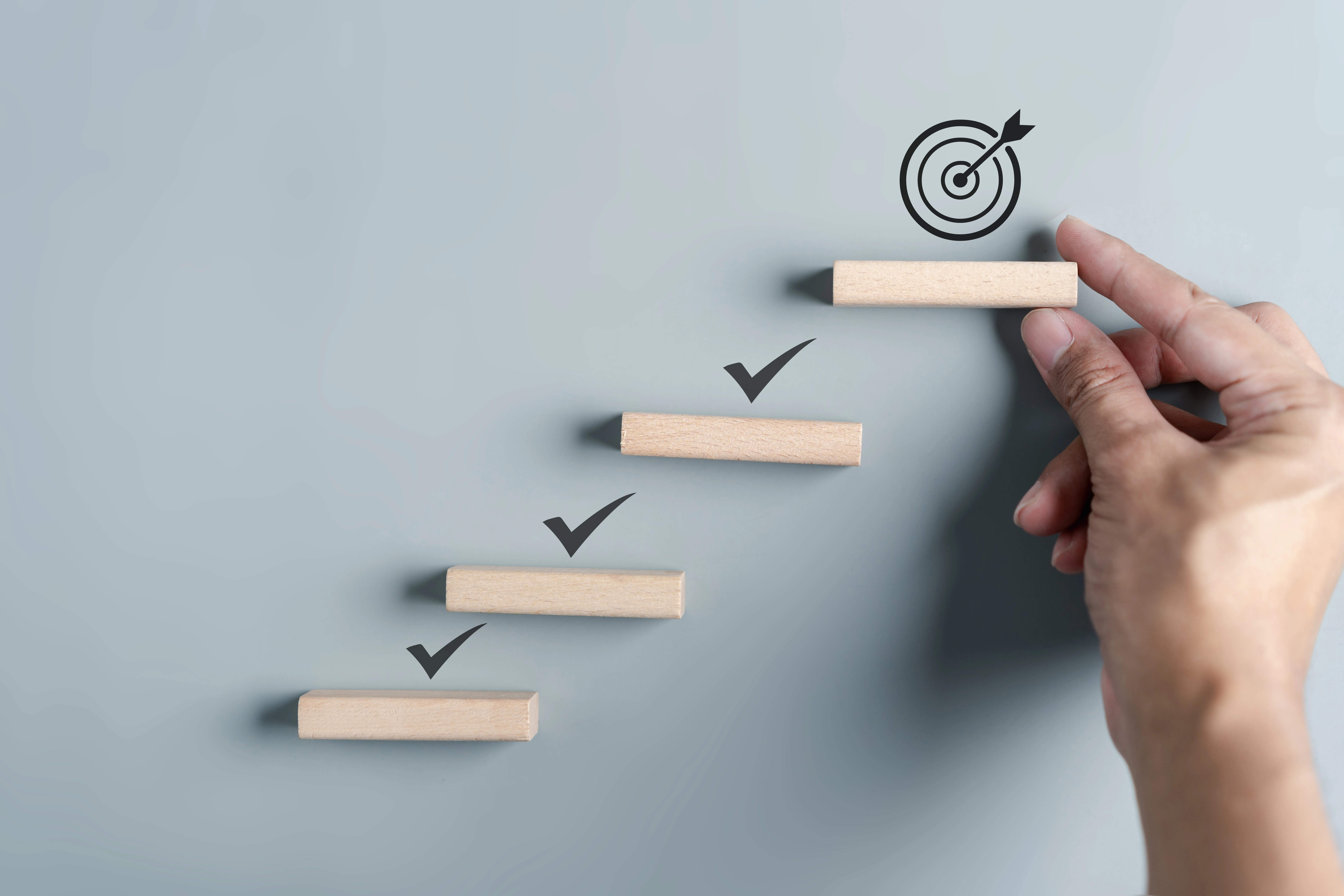 Hand placing the final wooden block on a staircase-like arrangement with checkmarks, leading to a target icon at the top.