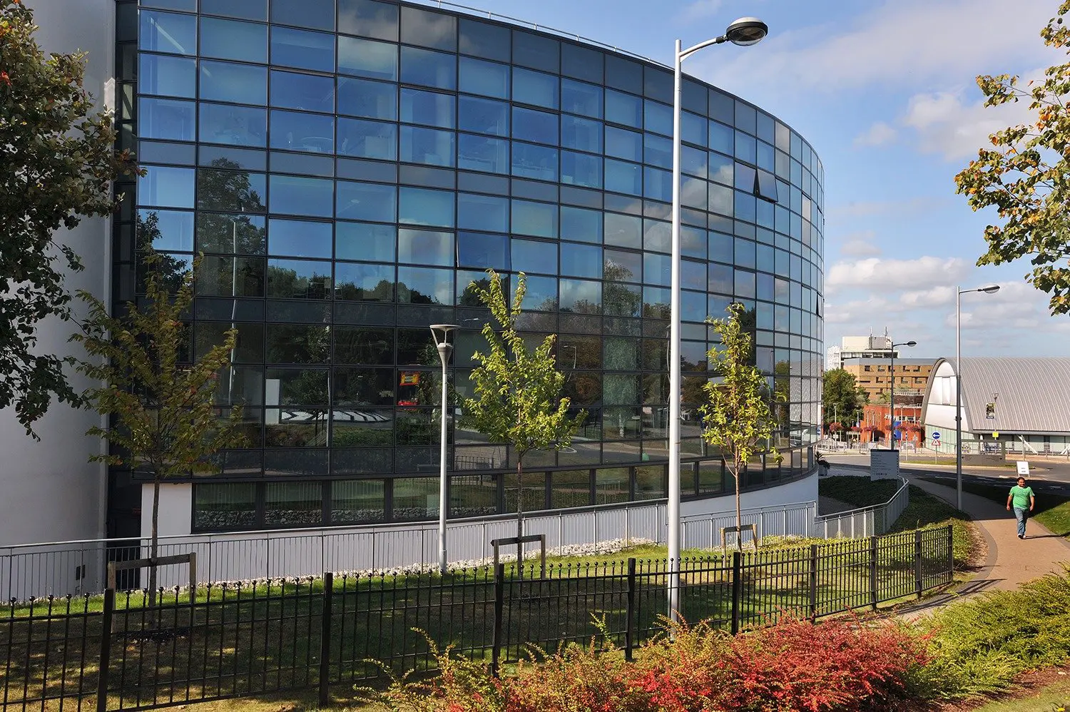 Modern glass-fronted university building with landscaped surroundings and pedestrian walkway in the sun.