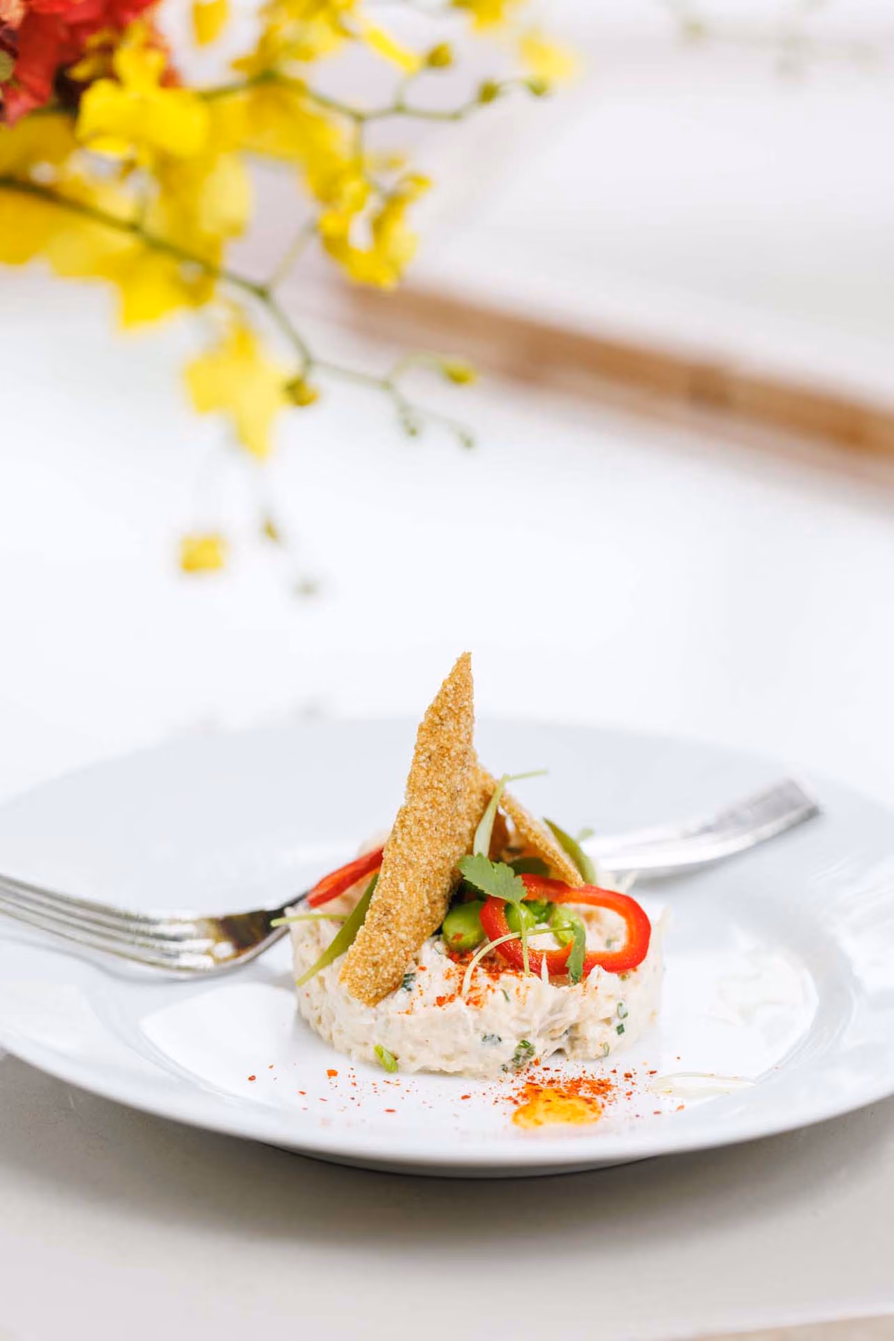Crab salad topped with a crisp shard and sliced chili peppers beside yellow orchids