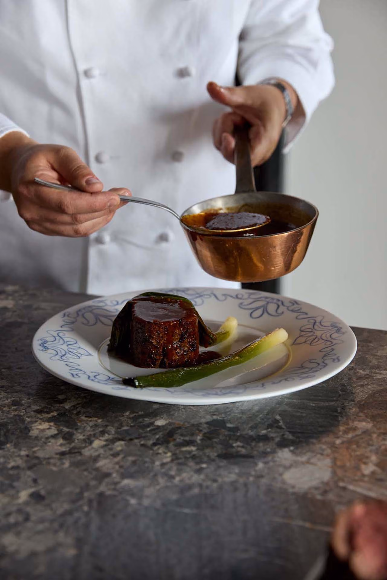 Chef spooning sauce over a beef fillet with charred scallion on a blue-patterned plate