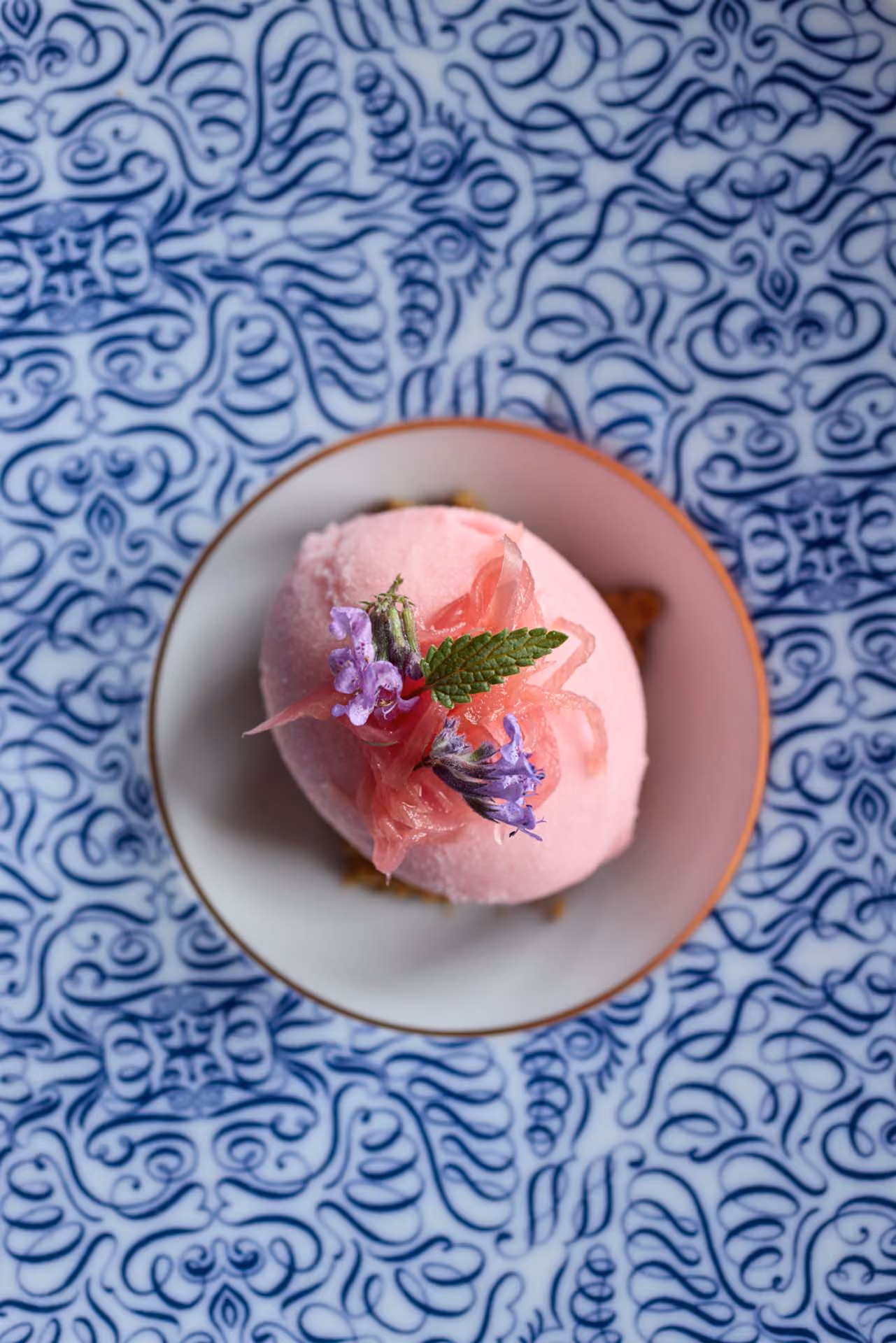 Overhead pink sorbet scoop garnished with mint and purple flowers on a patterned blue plate