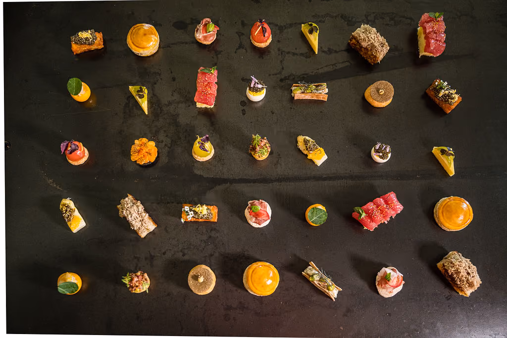 Overhead grid of assorted plated canapes arranged in rows on a dark table