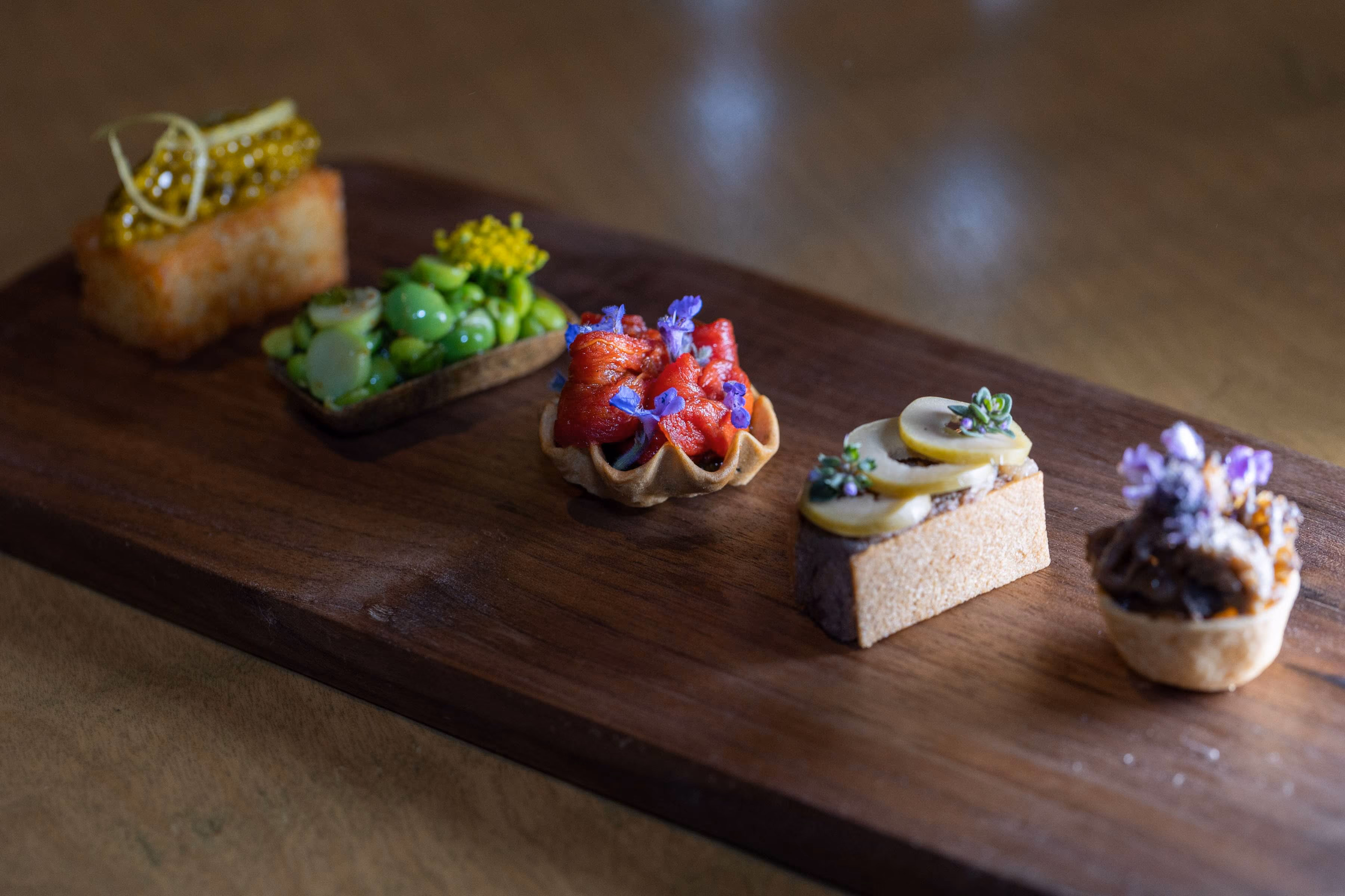 Assorted canapes on a wooden board including caviar toast, edamame, tomato tart, and mushroom tartlet
