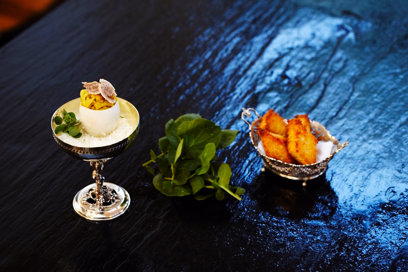 Elegant appetizer trio on a slate board: a truffle-topped egg shell cup, watercress, and fried bites in a silver basket