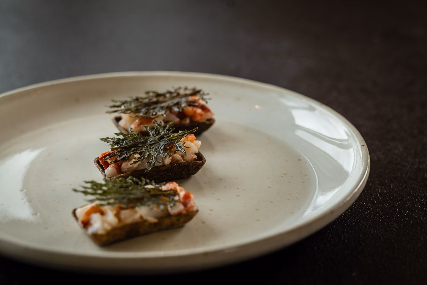 Three seaweed-topped seafood canapes on a dark cracker base