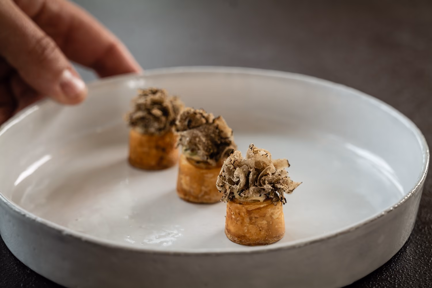 Hand placing shaved truffle-topped puff pastry bites on a white dish
