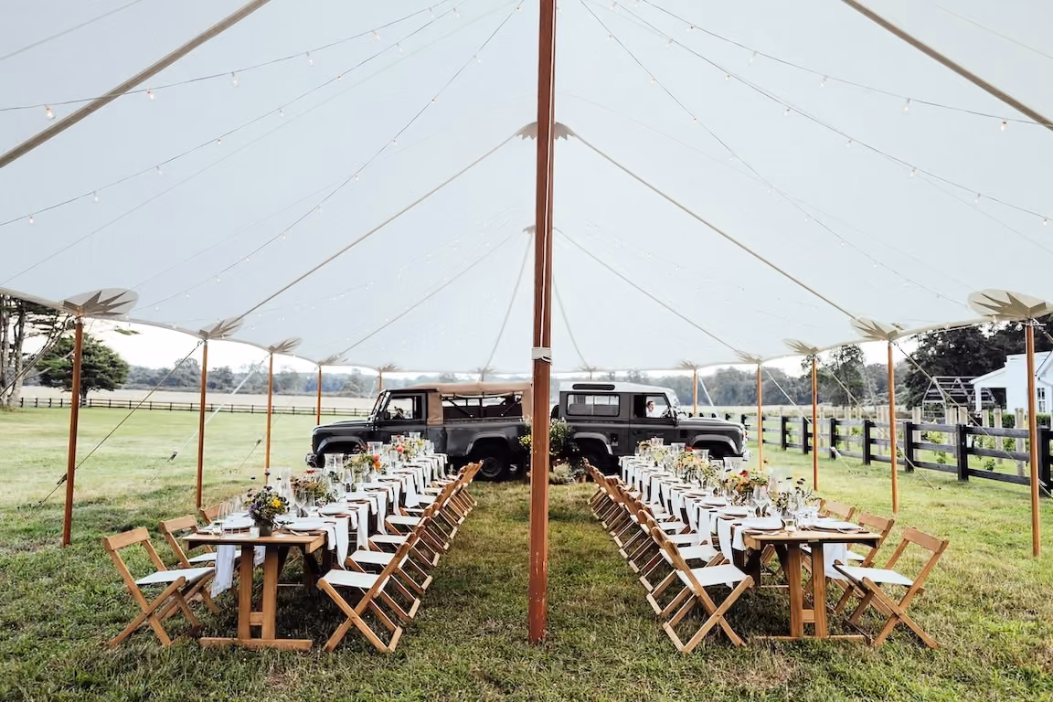 Long outdoor dinner tables set under a tent with two vintage Land Rovers parked behind
