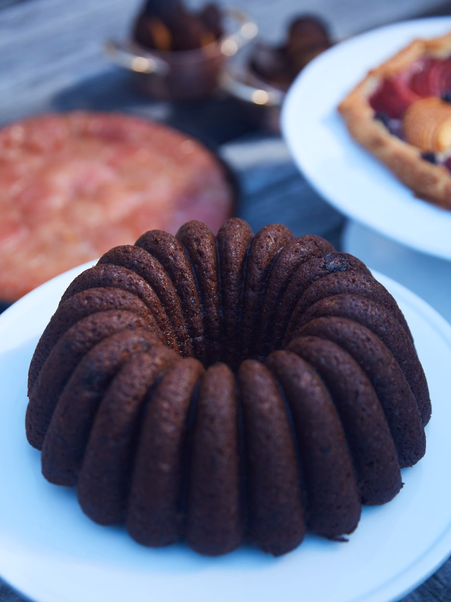 Chocolate bundt cake on a plate with other desserts blurred in the background