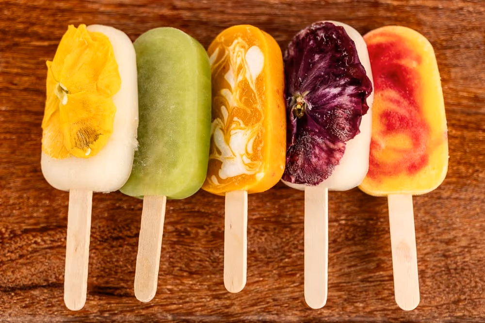 Five popsicles topped with edible flowers and fruit swirls on a wooden board