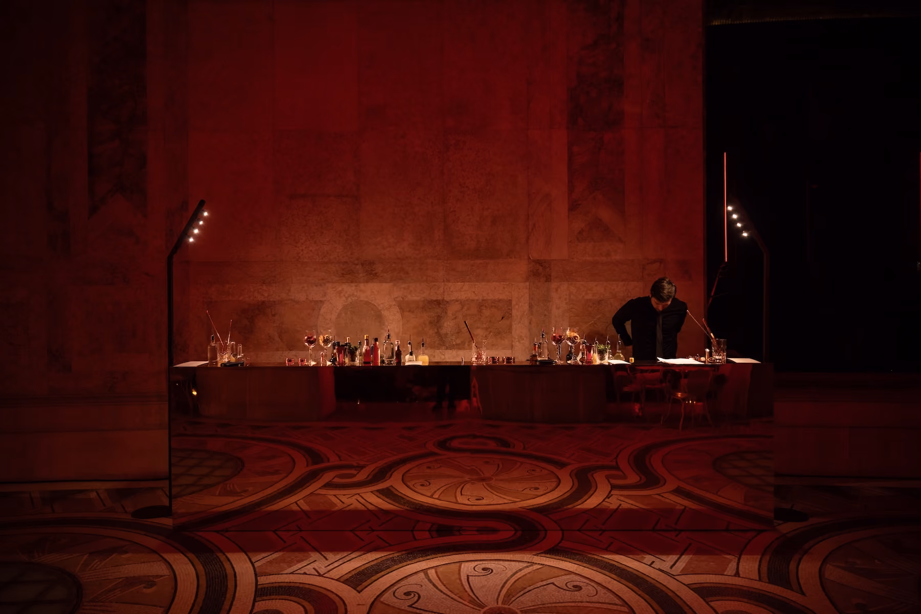Red-lit bar setup in an ornate hall with a bartender preparing drinks